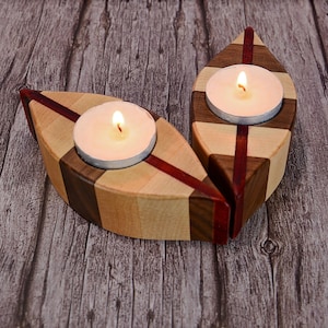 May include: Two wooden candle holders with a leaf shape design. Each holder has a small, round, white candle inside. The holders are made of different types of wood, including walnut, maple, and cherry. The wood is stained in a natural color.