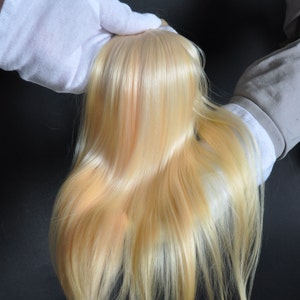 May include: A bundle of long, straight, light blonde human hair extensions. The hair is held in place by two gloved hands.
