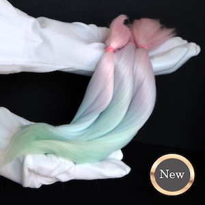Peut inclure: Trois mèches d'extensions de cheveux ombrées dans des tons de bleu clair, de vert et de rose. Les cheveux sont tenus dans une paire de mains gantées de blanc. L'image a un cercle doré avec le mot "Nouveau" en texte noir.