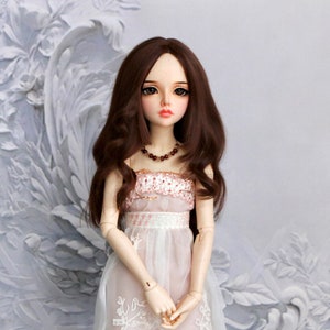 Puede incluir: Una muñeca de porcelana con el pelo largo y castaño que lleva un vestido blanco con detalles de encaje y un corpiño rosa. La muñeca tiene ojos marrones y lleva un collar.