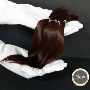 Puede incluir: Varias hebras de extensiones de cabello humano marrón oscuro atadas con gomas elásticas de colores. El cabello se sostiene en un par de guantes blancos sobre un fondo negro.