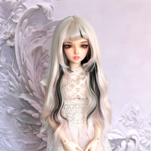 Puede incluir: Una muñeca con el pelo largo, blanco y negro, que lleva un vestido blanco de encaje. La muñeca tiene una tez rosa claro y ojos azules.