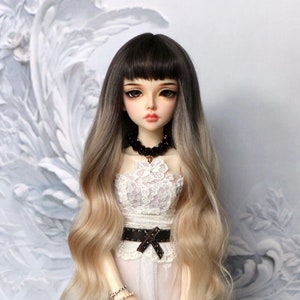 Puede incluir: Una muñeca con el pelo largo y degradado en tonos marrones y rubios, que lleva un vestido blanco de encaje con un cinturón negro y un collar de cuentas negras.