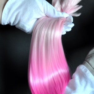 Puede incluir: Una larga hebra de cabello sintético con un degradado de color de blanco a rosa. El cabello se sostiene en dos manos enguantadas de blanco.
