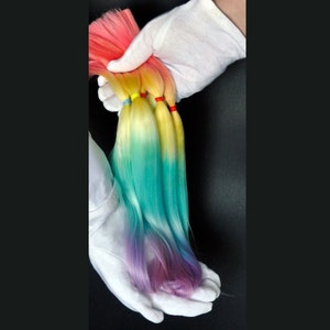 Puede incluir: Tres mechones de extensiones de cabello ombré en tonos rosa, amarillo, azul y morado. El cabello está atado con gomas elásticas de colores.