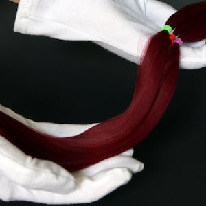 Puede incluir: Dos manos enguantadas de blanco sujetan una larga hebra de extensiones de cabello humano de color rojo oscuro.