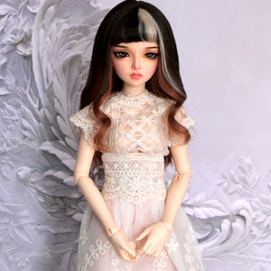 Puede incluir: Una muñeca con el pelo largo y ondulado marrón y blanco, que lleva un vestido blanco de encaje con cuello alto y mangas cortas.