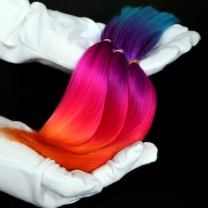 Puede incluir: Tres mechones de extensiones de cabello sintético en tonos naranja, rosa, morado y azul. El cabello se sostiene en un par de guantes blancos.