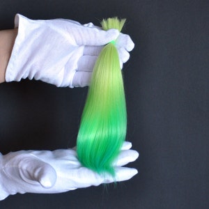 Puede incluir: Una sola hebra de cabello sintético en un degradado de verde claro a verde oscuro. El cabello se sostiene en un par de guantes de algodón blanco.