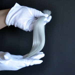 Puede incluir: Una sola hebra de extensión de cabello humano gris plateado sostenida en dos manos enguantadas.