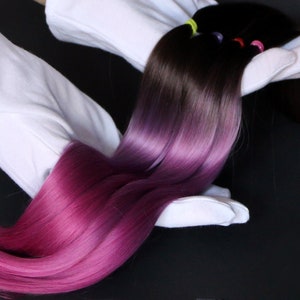 Puede incluir: Una larga hebra de extensiones de cabello humano con un degradado de color de marrón oscuro a morado. El cabello se muestra en guantes blancos.
