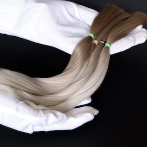 May include: Three strands of human hair extensions, one blonde, one light brown, and one dark brown, are held in white gloves. The hair is tied with colorful rubber bands.