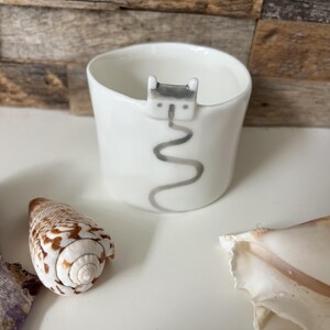 May include: A white ceramic cup featuring a small, sculpted grey cat figure perched on the rim, with a painted grey wavy line extending downwards from the cat along the exterior surface of the cup, accompanied by two seashells nearby.