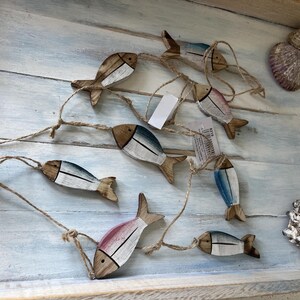 May include: A decorative garland featuring seven small wooden fish shapes connected by natural jute twine. Each fish is painted with white and blue or pink accents, showing a distressed finish. Two seashells are placed near the wooden fish.
