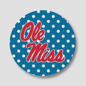 May include: A circular sticker featuring a blue background with a white polka dot pattern. The text Ole Miss is printed in the center in a red, cursive script font with a white outline, creating a bold and clear visual design for this item.