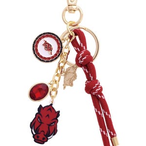 May include: Gold-tone keychain with a red and white striped rope, a red jewel, and a red and white medallion. The keychain features a red and white Razorback mascot charm and a gold-tone chain with a small charm.