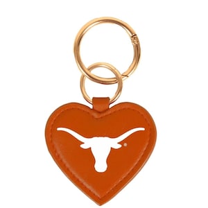 May include: A heart-shaped keychain in burnt orange leather features a white longhorn logo. The keychain has a gold-colored metal ring and clasp for attaching to keys or a bag. The design is simple and classic.