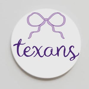 May include: A white, round coaster with a purple bow illustration at the top and the word "texans" in a cursive font below. The coaster is a simple, yet stylish design.