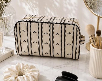 May include: A rectangular toiletry bag featuring a cream-colored base with vertical black stripes and small black floral motifs. The bag has a gold-toned zipper closure along the top edge and a small fabric tab on the side for easy handling.