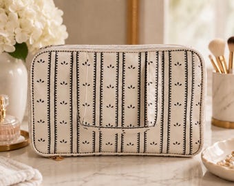 May include: A rectangular zippered travel case in an off-white color featuring a repeating pattern of vertical black stripes and small black floral motifs. The bag includes a top handle and a gold-toned zipper pull, with a structured, boxy shape.
