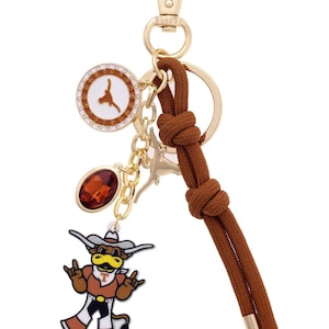 May include: A gold-toned keychain with a brown rope detail and multiple charms. Charms include a round emblem with a Longhorn logo, a faceted orange gem, and a cartoon Longhorn mascot. The keychain has a lobster clasp.