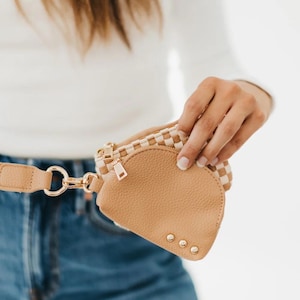 May include: Tan leather fanny pack with a gold zipper and gold studs. The top section has a woven checkered pattern. The adjustable belt has a gold clasp. The fanny pack is being held by a person.