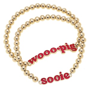 May include: Two gold beaded bracelets with red enamel text that reads "woo-pig" and "soooie". The bracelets are on a white background. The beads are small and round.
