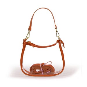 May include: A clear, transparent handbag with a brown handle and strap. The bag has a rounded shape with brown trim and gold-colored hardware. The detachable strap is rolled up inside the bag.