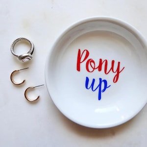 May include: A white ceramic trinket dish with the words "Pony up" in red and blue script.  Also, a silver ring with a textured band and a clear stone, and two gold hoop earrings are next to the dish.