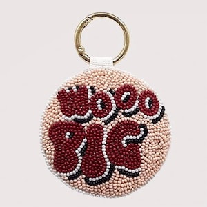 May include: A round beaded keychain with the words "WOO BIG" in burgundy, black, and white beads. The background is a light tan color, and the keychain has a gold-colored ring.