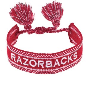 May include: Red and white woven bracelet with the word "RAZORBACKS" embroidered in white. The bracelet has two red tassels with white accents. A stylish accessory for fans.