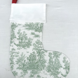May include: A Christmas stocking with a white cuff and a red hanging loop. The body of the stocking features a green and white toile pattern depicting pastoral scenes with figures, trees, and foliage. The stocking is made of fabric.