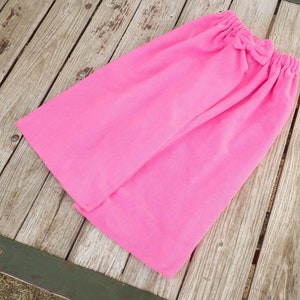 May include: A bright pink skirt with an elastic waistband and a decorative bow. The skirt is made of a soft fabric and is laid out on a weathered wooden surface. The skirt is a solid color.