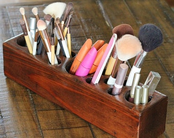 May include: A wooden makeup brush holder with multiple compartments, filled with various makeup brushes, lipsticks, and other makeup tools. The holder is stained a dark brown color and has a rustic look.
