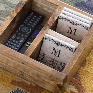 May include: A wooden box with a rustic walnut finish, divided into compartments. The box contains a remote control and a stack of coasters with a black and white floral design and the letter "M" in the center. The box is labeled "GREY RIVER Market".