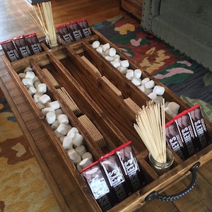 May include: Wooden tray with dividers for holding s'mores ingredients. The tray has two long compartments for graham crackers and marshmallows, a smaller compartment for chocolate bars, and a small square compartment for skewers. The tray has a black metal handle.