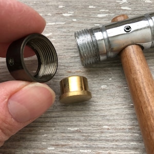 May include: A close-up of a metal hammer with a wooden handle, a brass ring, and a black metal ring. The hammer is used for jewellery making.