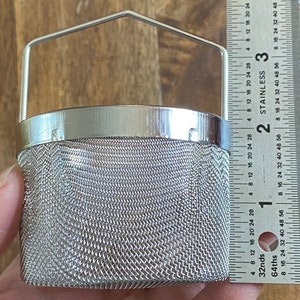 May include: A silver mesh tea infuser with a handle. The infuser is about 2 inches in diameter and 3 inches tall. The infuser is sitting on a wooden surface.
