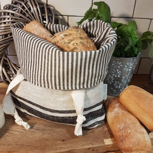 May include: A bread basket with black and white stripes, containing two loaves of bread. The basket has a drawstring closure and is on a wooden surface. Two more loaves of bread are placed beside the basket.
