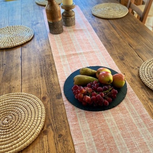 Op de afbeelding: Een houten eettafel met een perzik- en wit gestreepte tafelloper, kaarsen en geweven placemats. Een zwarte schaal bevat druiven, peren en appels.