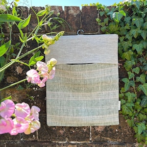 May include: A two-tone linen wall pocket, hanging on a brick wall. The top section is plain light grey, the lower section has horizontal stripes in light green and beige. The pocket hangs from a metal ring.