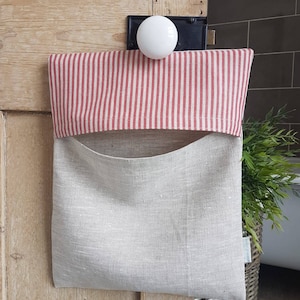 Könnte beinhalten: Ein Leinen-Taschen-Organizer mit rot-weiß gestreiftem Oberteil, der an einer Holztür hängt. Die Tasche hat eine natürliche Leinenfarbe mit einer Vordertasche. Ein weißer Türknauf befindet sich darüber.