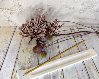 May include: A white ceramic incense holder with a rectangular design. A lit incense stick rests in the holder. Dried floral arrangement with pink and green accents is in the background. The holder is on a wooden surface.
