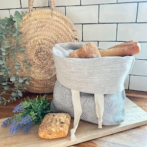May include: A bread basket with a grey and white patterned base and a light grey linen top, filled with loaves of bread. A round woven basket and sprigs of lavender are in the background. The basket has cream-coloured ties.