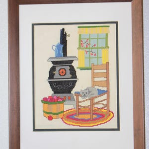 May include: Cross-stitch artwork depicting a cozy interior scene. A black stove with an orange design sits next to a wooden bucket filled with red apples. A gray cat rests on a wooden chair, and a colorful rug is on the floor. A blue pitcher is on top of the stove.
