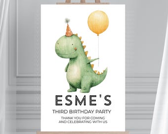 May include: A white sign on a wooden easel features a watercolor illustration of a green dinosaur wearing a party hat and holding a yellow balloon. Text reads "Esme's Third Birthday Party" and a thank you message.