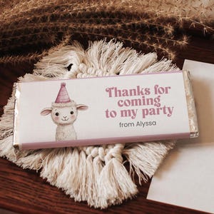Può includere: Un involucro di barretta di cioccolato rosa e bianco con un agnello di cartone animato che indossa un cappello da festa rosa. L'involucro dice "Grazie per essere venuto alla mia festa da Alyssa".