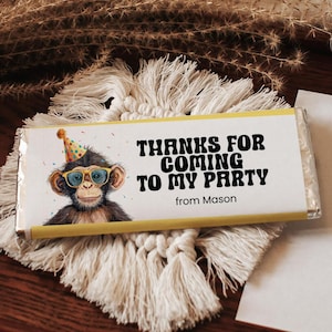 May include: A chocolate bar wrapper with a cartoon monkey wearing glasses and a party hat. The text reads "THANKS FOR COMING TO MY PARTY" and "from Mason". The wrapper has a yellow border and is on a macrame coaster.
