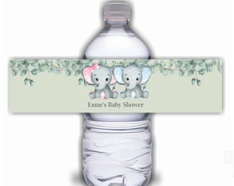 May include: Water bottle label with a green background and a eucalyptus garland. Two gray elephants with pink and blue accents are sitting on the garland. The text "Esmé's Baby Shower" is printed below the elephants.