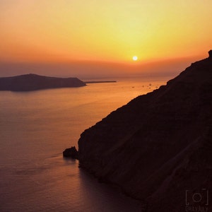 May include: A scenic sunset over a body of water with a large, dark cliff in the foreground. The sun is setting behind a distant island, casting a warm orange glow over the scene.
